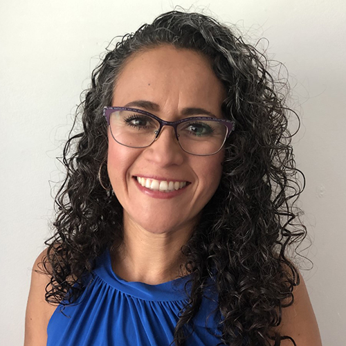 portrait of Erika Cedillo: woman with long curly grey and black hair smiling. she's wearing hoop earrings and purple rimmed glasses.