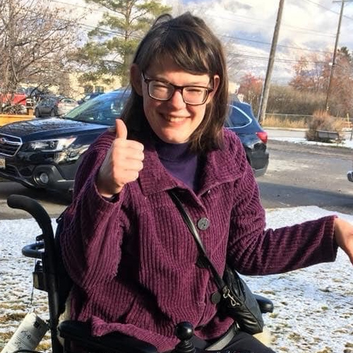 woman with long dark brown hair and large brown glasses in a wheelchair giving a thumbs up while smiling