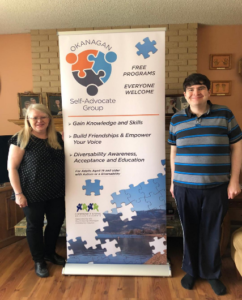 A woman with long blonde hair and a taller man with short black hair stand on either side of a vertical banner for the Oakanagan Self-Advocate Group. It reads: Oakanagan Self-Advocate Group. Free programs. Everyone welcome. Gain knowledge and skills. Build Friendships and empower your voice. Diversability awareness, acceptance, and education.