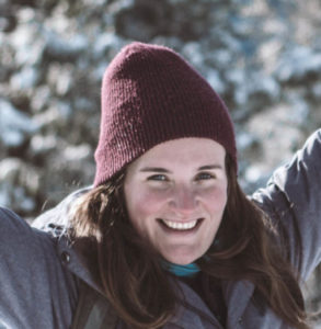 a woman with long brown hair standing in the snow-covered wilderness at a high point with the view of trees and mountains under a blue sky behind her. She is wearing a red toque and a grey jacket and has her arms outstretched and is smiling while looking at the camera.