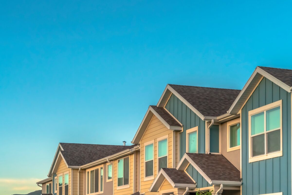 A clear blue sky above a the roofs of a row of connected townhomes. Some of the exterior of the houses are a light yellow and some a deep blue.