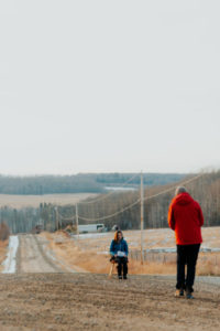 A long dirt road visible to the horizon. A young woman is sitting on a chair in the middle of the road looking down at some paper in her hand. A person in a red jacket is standing facing her with their back facing the camera. There is a light dusting of snow on the edges of the road and surrounding hilly land. 