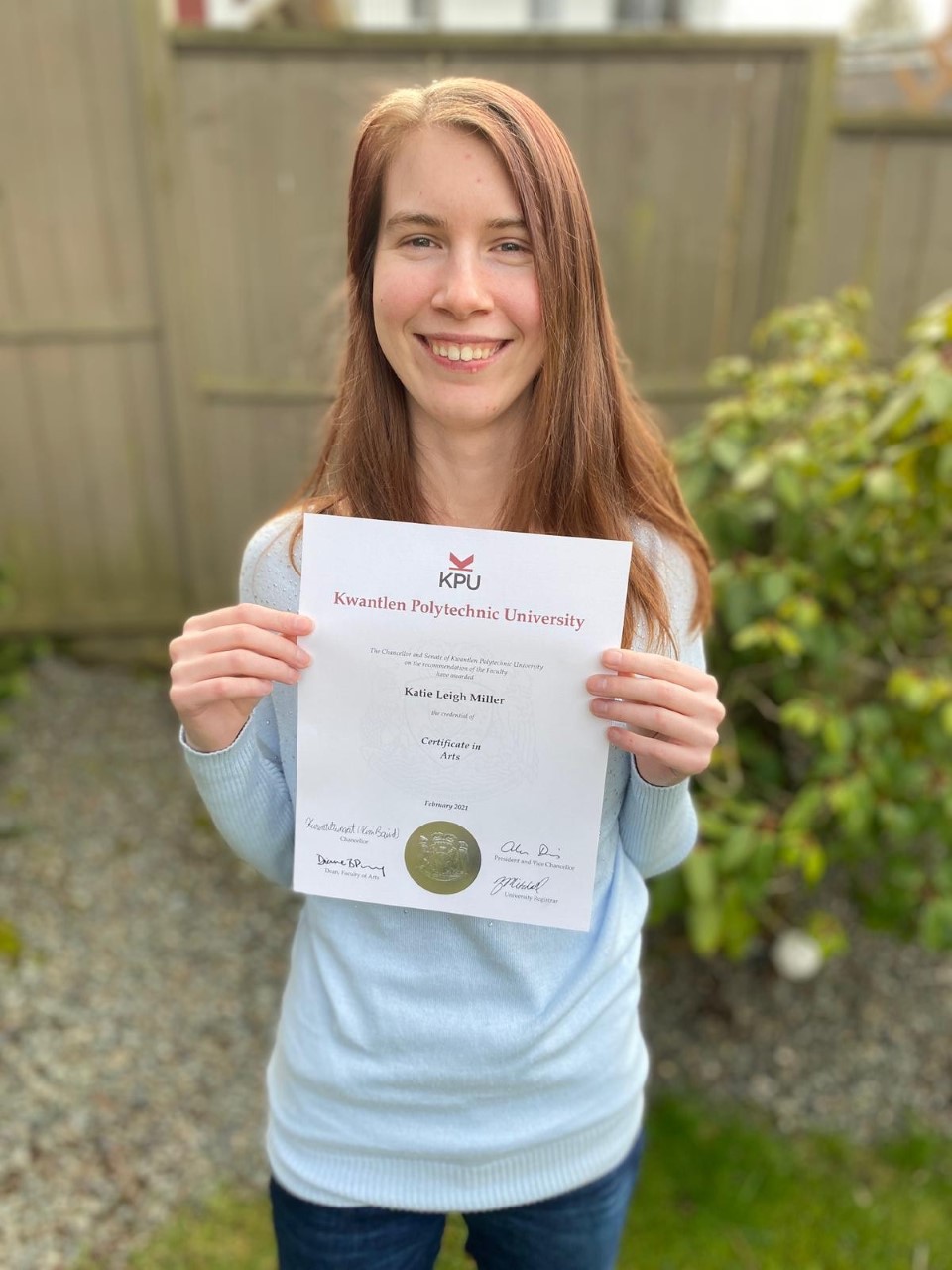a young woman with long brown hair is smiling at the camera while holding a certificate from Kwantlen Polytechnic University. It shows her name in black text as Katie Leigh Miller. Text below that reads Certificate in Arts. there are signatures and a large golden seal at the bottom of the certificate. Katie is wearing a light blue sweater and dark blue jeans. She is standing in what appears to be a back yard with a tall wooden fence behind her.