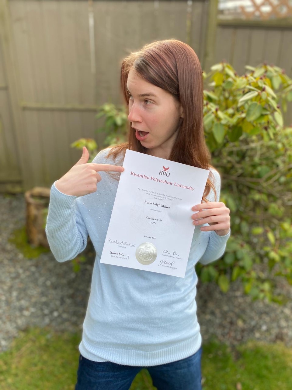 a young woman with long brown hair holds a certificate from Kwantlen Polytechnic University while pointing at it with one hand. She is looking off to the side and is making an expression of surprise. It shows her name in black text as Katie Leigh Miller. Text below that reads Certificate in Arts. there are signatures and a large golden seal at the bottom of the certificate. Katie is wearing a light blue sweater and dark blue jeans. She is standing in what appears to be a back yard with a tall wooden fence behind her.