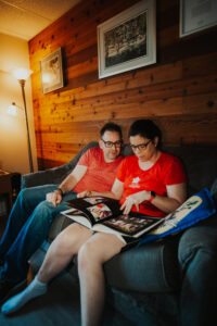 A man an woman seated on a couch in a room with dim lighting and a wooden wall behind. They are wearing red tshirts and have glasses on. They are looking through a large photo album together