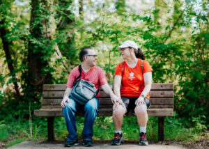 A man and woman, both wearing red tshirts sitting on a bench in a park with tall trees in the background. The man has a grey shoulder bag on. They are both wearing glasses