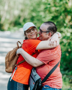A man and woman both wearing red shirts hugging outside with greenery in the background. The woman has a leather backpack. The man has a grey shoulder bag. They are both wearing glasses