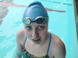 A young woman standing in a pool. She is wearing a light blue swimming cap and dark-tinted goggles propped up on her forehead. 
