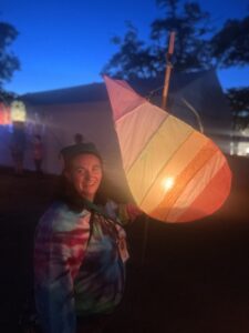 Alison Klein. a woman with wavy brown hair wearing a tie die rainbow sweater smiling. She's outside at night where glowing rainbow lanterns are.