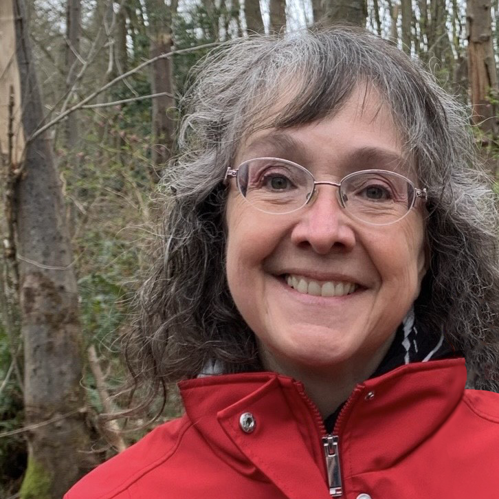 A woman with long wavy grey hair. She is wearing round rimless glasses and a red jacket. She is smiling at the camera. her shoulders and head are visible. The background is a forest on a cloudy day.