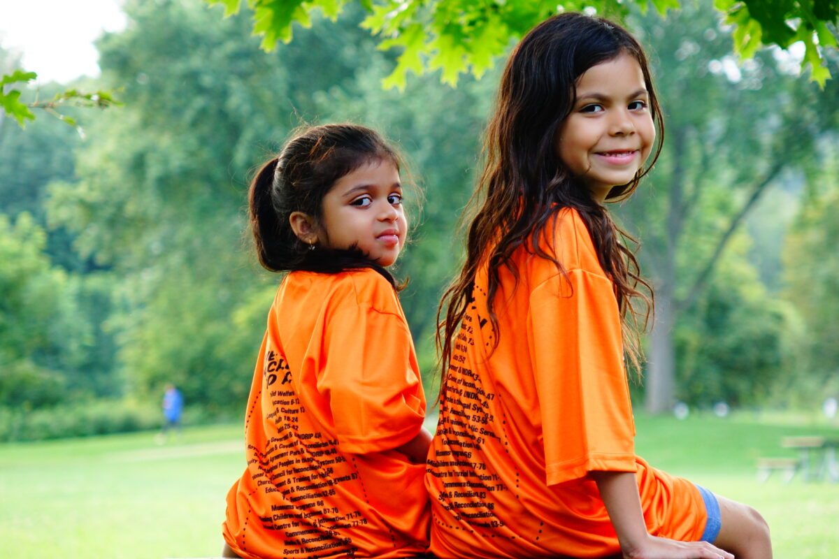 two young girls with long dark brown hair sitting outside. They are facing away from the camera and have their heads turned towards the camera. They are smiling. They are wearing orange shirts with words all over the back for national day for truth and reconciliation.