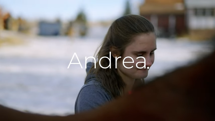 A young woman with long brown hair stands outside. She is looking down at something she is doing and is visible from the shoulder up. White text over the image reads: Andrea.