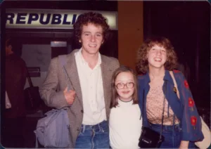 a young man, and a young woman stand on either side of a young girl who is wearing round glasses. The photo looks older from the 70s.