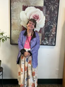 An elderly woman with short brown hair stands in front of a large painting of a white rose. She is wearing glasses, and a purple jacket over a pink shirt