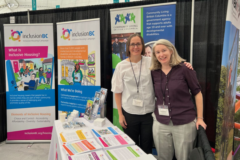 Two women standing at an exhibit table with colourful papers on it. Behind them are two brightly colored banners with facts on inclusive housing on them and illustrations of diverse communities