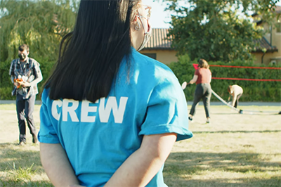 A person with long black hair seen from behind standing outside in a sports field. They are wearing a light blue shirt that has text on the back that reads: Crew