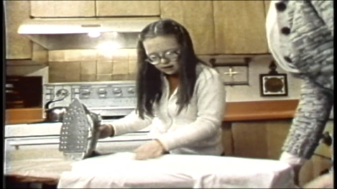 a young girl with round glasses holds an iron over and ironing board with a white cloth on it. The photo looks old and she is in a kitchen with a stove visible in the background