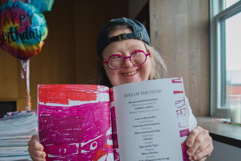 A woman wearing a baseball cap and pink-rimmed round glasses holding up a book opening it for the camera.