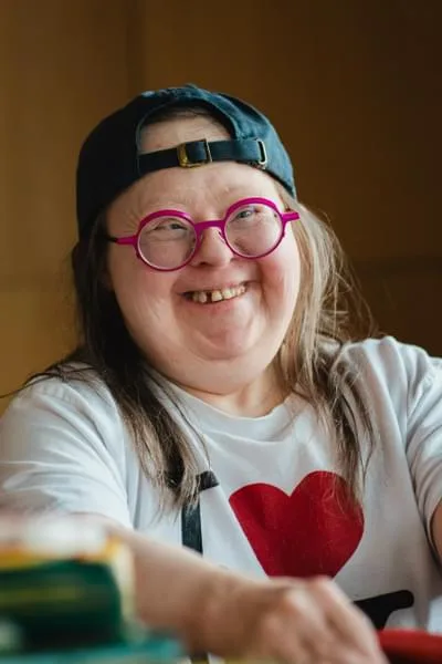 A woman wearing a baseball cap and pink-rimmed round glasses smiling. She is wearing a white shirt with a red heart on it and black text.