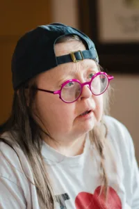 A woman wearing a baseball cap and pink-rimmed round glasses looking into the distance off camera