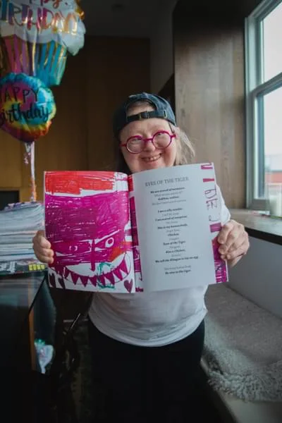 A woman wearing a baseball cap and pink-rimmed round glasses holding up a book opening it for the camera.