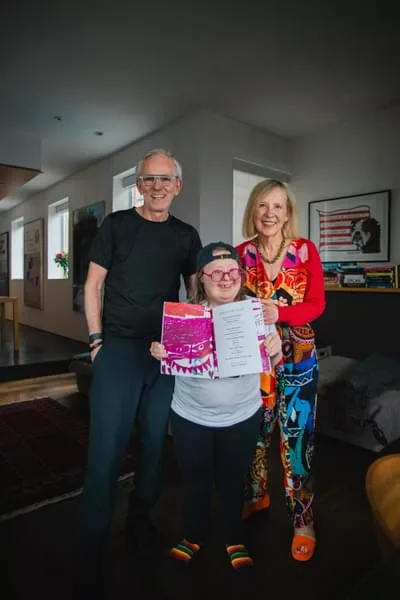 A woman wearing a baseball cap and pink-rimmed round glasses holding up a book opening it for the camera. A man and a woman stand to either side of her smiling