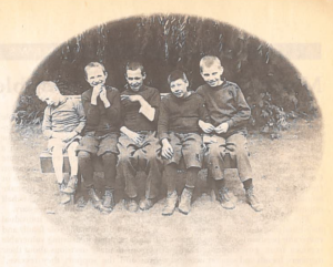 A black and white image on newspaper. There are five young boys with short hair sitting in a row on a bench outside in front of trees. they are wearing sweaters and long pants. 