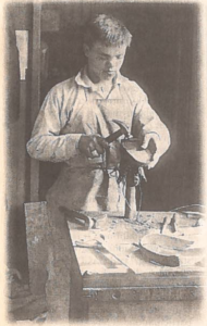 a black and white image on newspaper. A young man wearing a white shirt and an apron stands at a workshop table working on a shoe with a hammer in one hand. 