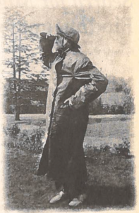 a black and white photo on newspaper. A person in a long trenchcoat stands outdoors with one hand held up to their forehead shielding their eyes from the sun. They are looking into the distance to the left of the image. 