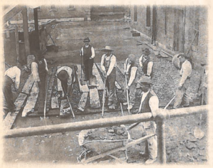 a black and white image on newspaper. several men in white shirts and dark vests with shovels on a building site working. one man is pushing a wheelbarrow in the foreground