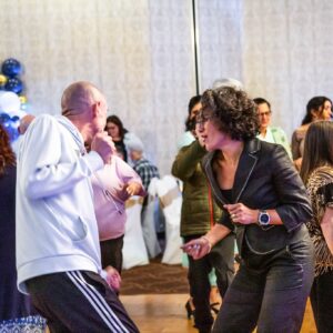 Erika Cedillo, a woman with long curly black hair, and Bryce Schaufelberger, a man with a bald hair and light skin, dance together on a dance floor within a large group of people generously spaced out. 