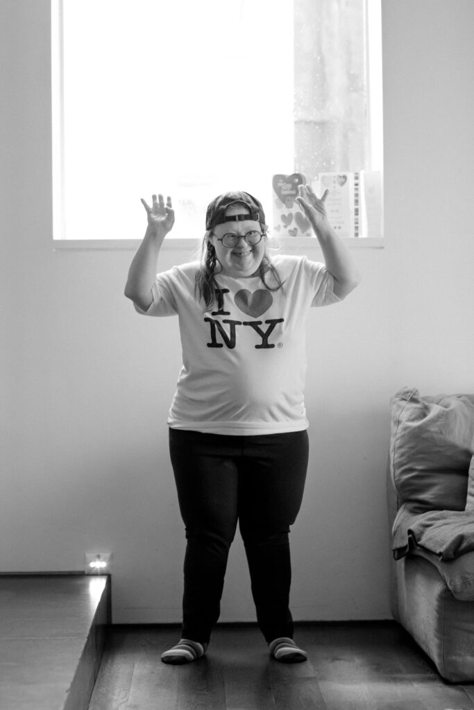A black and white photo of a woman standing in a room in front of a window. She has her hands up on the air and is smiling. She is wearing a baseball cap, round glasses, and a tshirt that says, I love N Y. with the word "love" being shown as the icon of a heart shape.