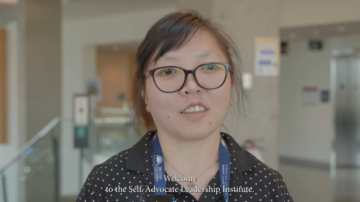 a young person with black rimmed glasses standing indoors. there are subtitles that read: Welcome to the Self Advocate Leadership Institute