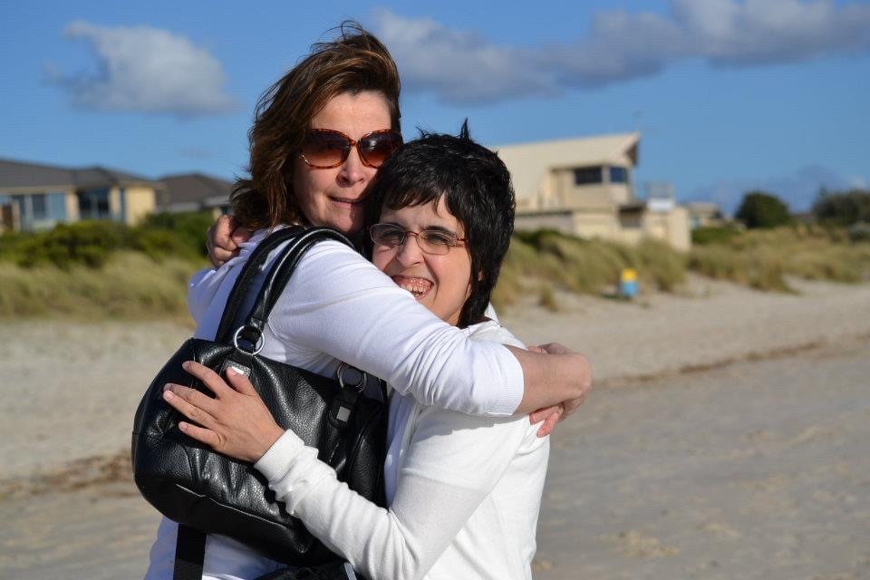 A mother and her adult daughter, both with dark long hair are hugging outside on a rocky beach while looking at the camera.
