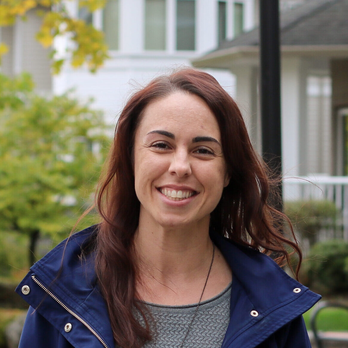 a woman with long brown hair outside wearing a grey shirt under a blue jacket. She is smiling. There are green trees and a white houses visible in the background