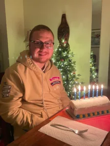 A young man with glasses and a light brown hooding sitting at a dining table with a cake in front of him. The cake has lit candles in a row on it. There is a christmas tree in the background.