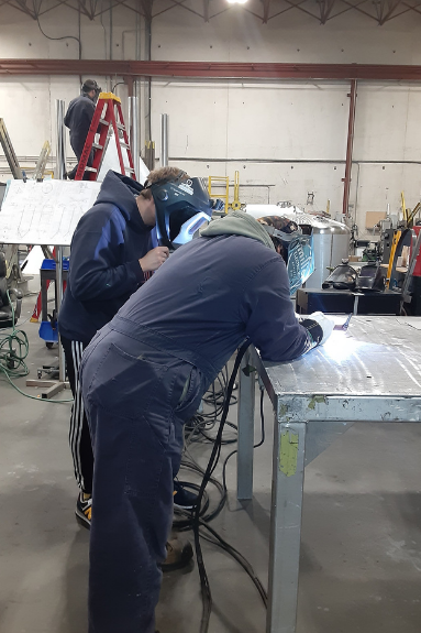 Two people at a large metal workbench with welding masks. They are looking closely at the top of the table and there is a bright light shining on both of them 