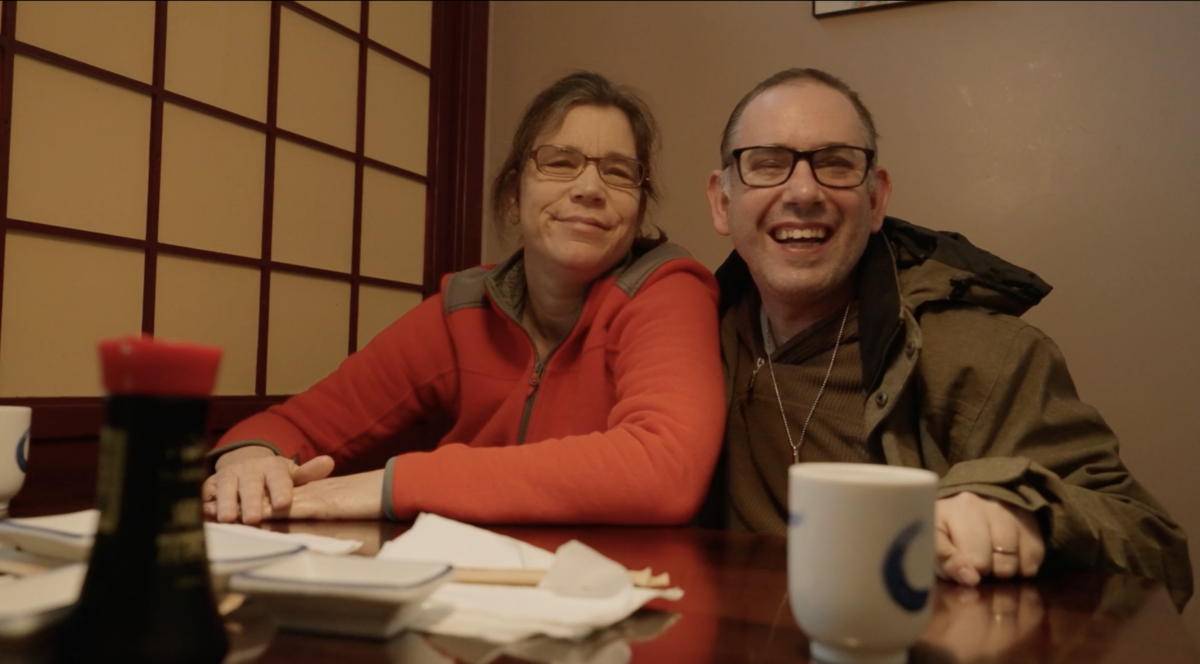 A couple seated on one side of a japanese restaurant table. They are a man and a woman and are wearing sweaters and are smiling at the camera.