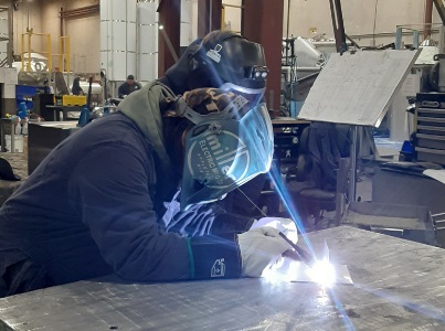 A person in a large industrial workshop wearing a welding mask and welding on the top of a large metal workbench. The welding is shining a bright light over the person's mask and body.