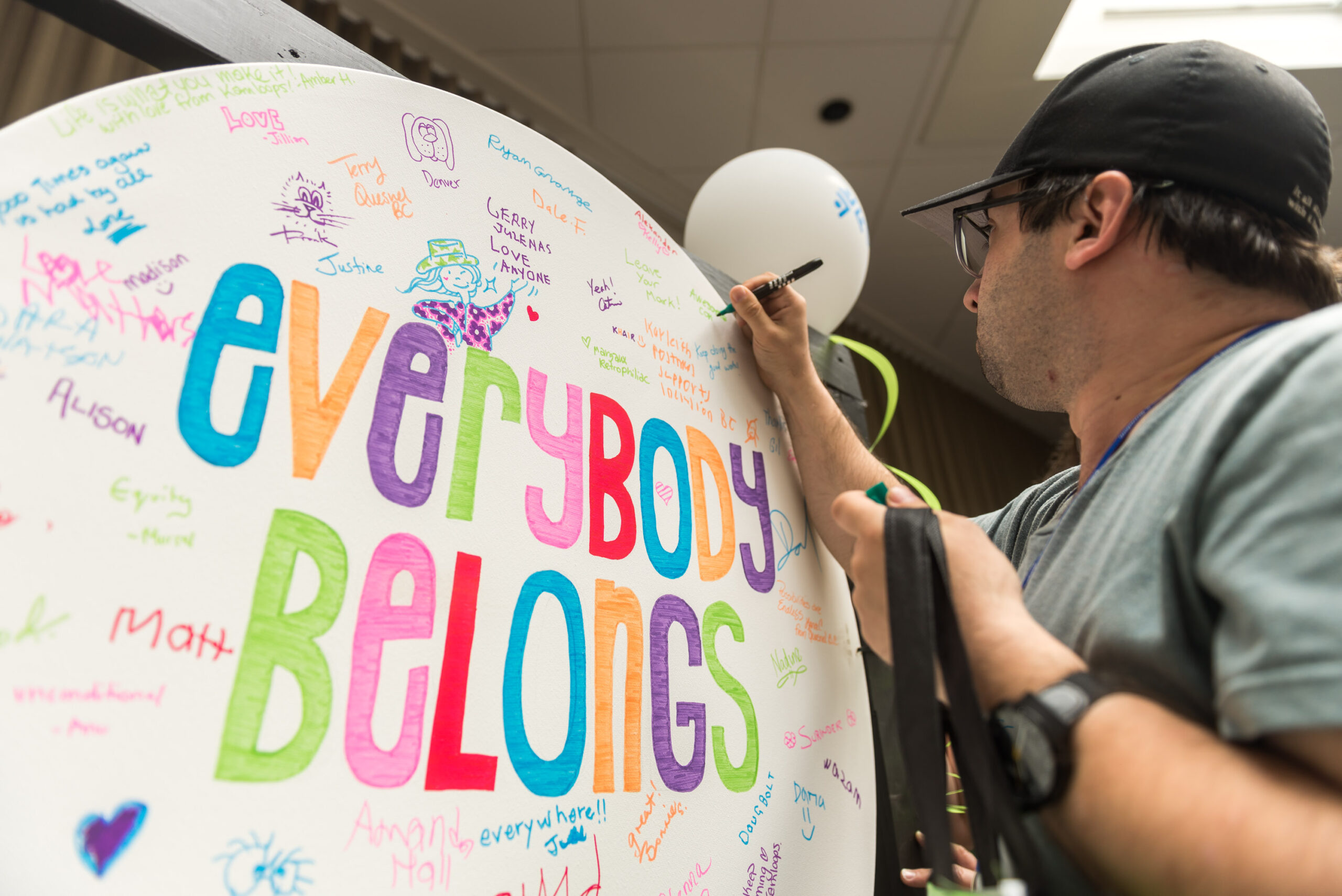 A person writing on a circular piece of paper that is upright. Everybody Belongs is written in large colourful letters. signatures and positive messages are written all over with colourful marker
