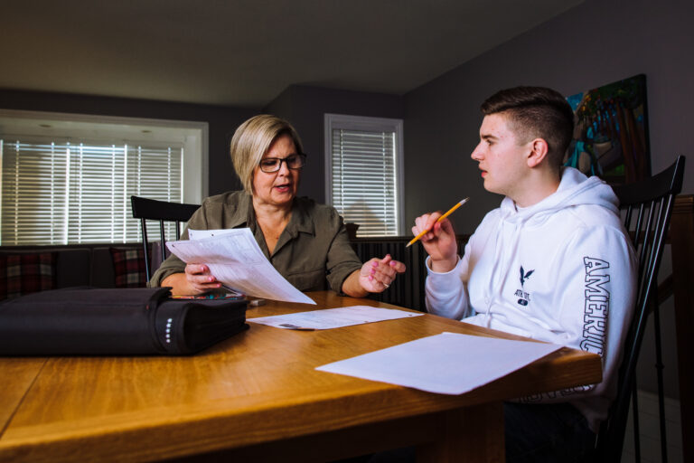 A woman and her teenage son sit at a dining table beside each other engaged in conversation while holding papers and pencils.