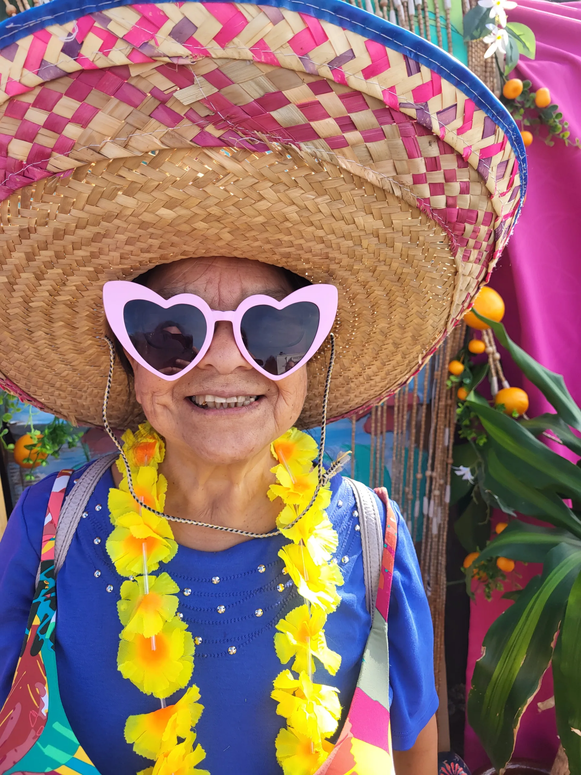 A person wearing a large straw hat with magenta weaved into it. The are wearing heart-shaped sunglasses and a yellow flower necklace.