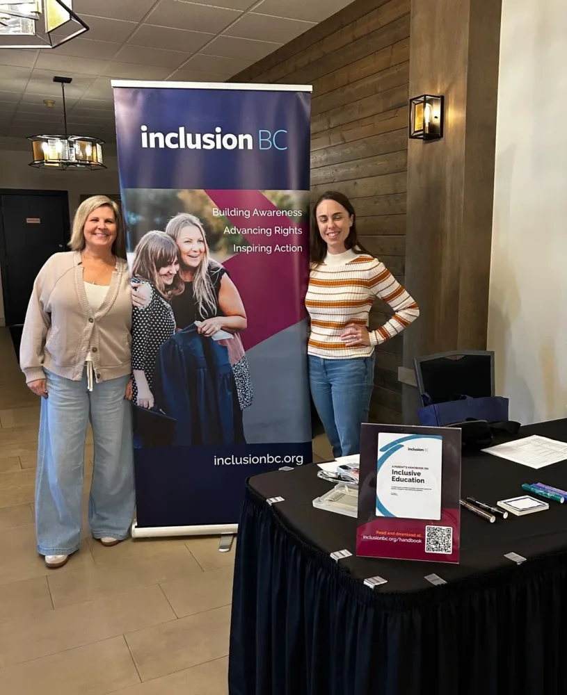 Two women standing beside an Inclusion BC banner at a registration table, smiling and ready to welcome attendees.