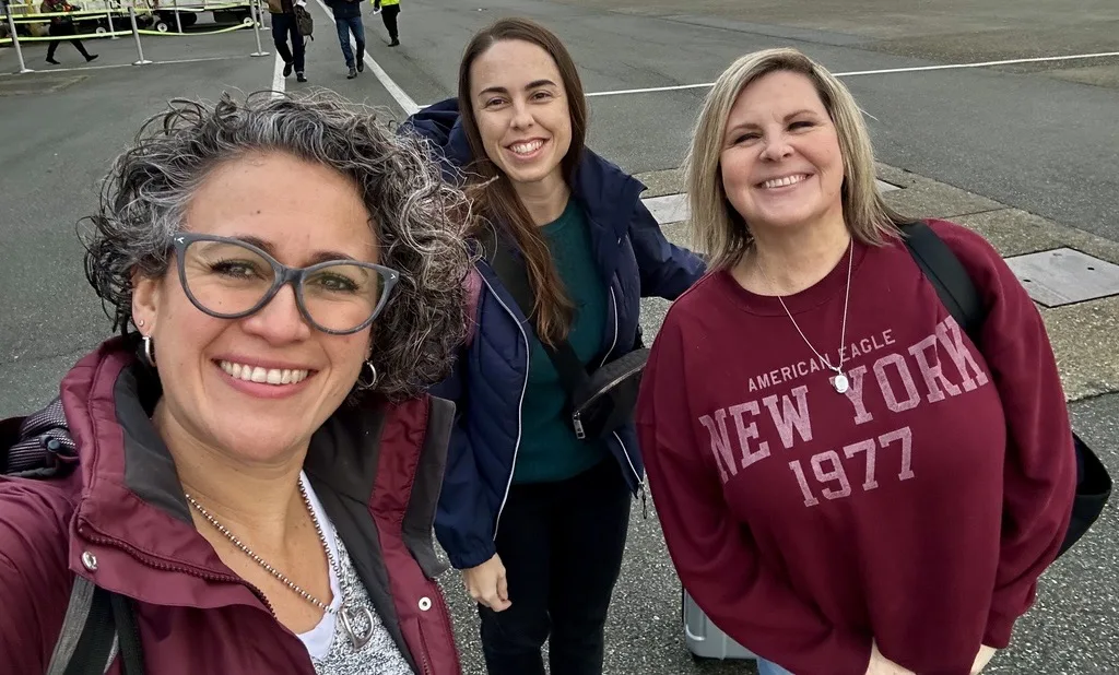 Three people are standing together outdoors an airport tarmac, dressed in casual jackets and sweaters. One person is taking the photo as a selfie, while luggage and airport personnel can be seen in the background.