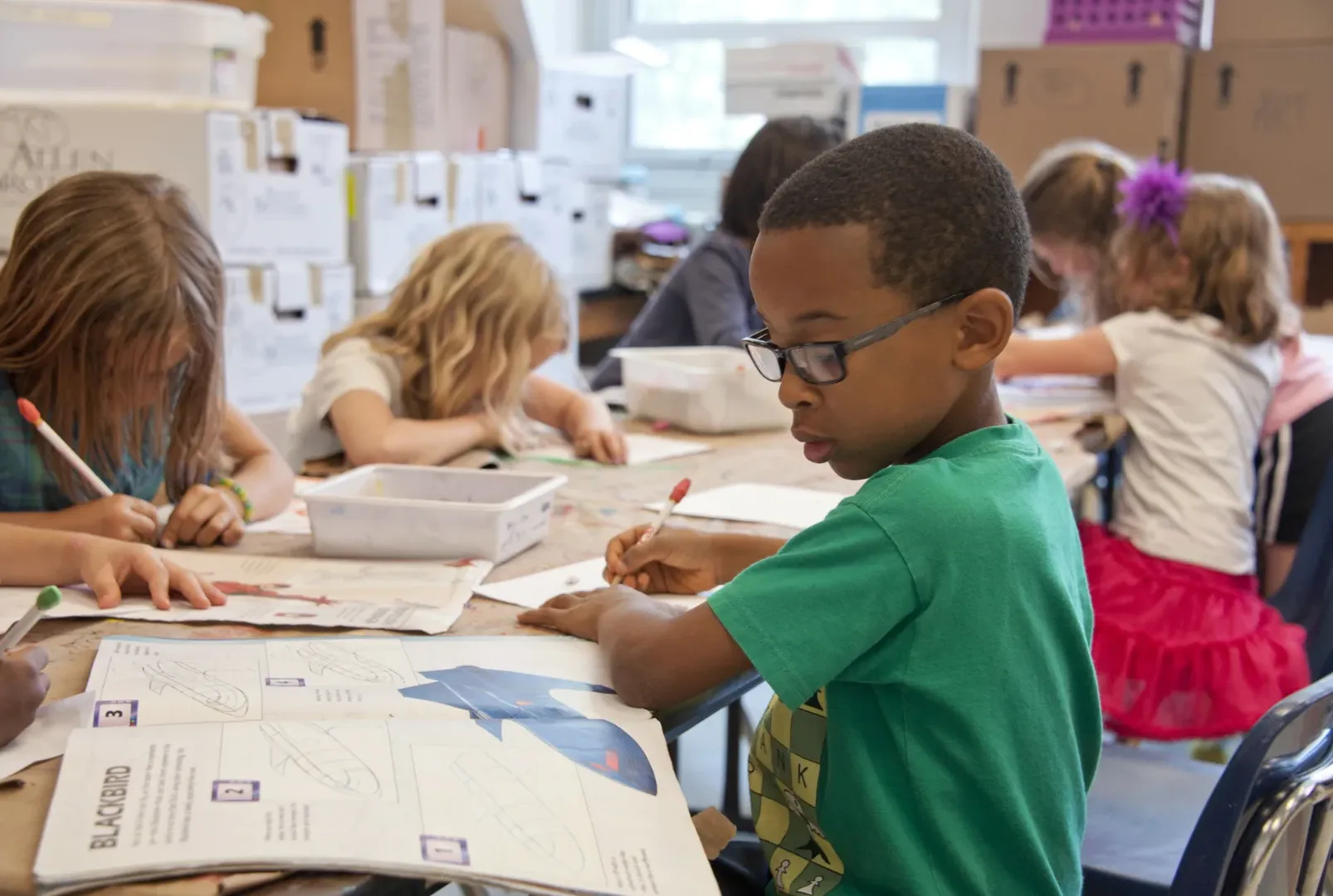 Children sit around a classroom table working on drawing and colouring activities. Art supplies, worksheets, and a booklet with an airplane diagram are spread across the table, and boxes and classroom materials are visible in the background.