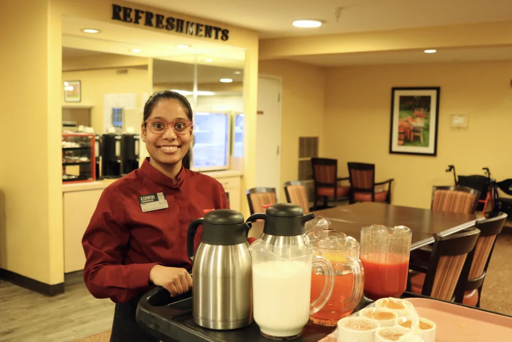 Staff member standing beside a refreshment cart with pitchers of drinks in a dining area.