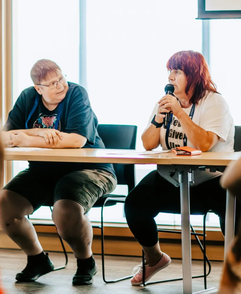 Two people sitting at a desk, one holding a mic.